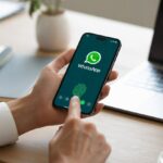 Pessoa segurando um smartphone com o aplicativo WhatsApp aberto, interagindo com o dispositivo em uma mesa de escritório organizada.