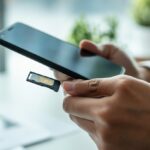 Pessoa segurando um smartphone com o compartimento do chip parcialmente aberto, em um ambiente de escritório claro.