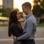 Um homem e uma mulher jovens estão juntos em um parque urbano ao pôr do sol, olhando um para o outro com carinho.