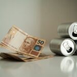Notas de cinquenta reais ao lado de latinhas de bebida sobre uma superfície limpa.
