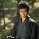 Um jovem guerreiro japonês com cabelo escuro e uma cicatriz na testa, vestindo um quimono verde e preto, em uma floresta com luz do sol filtrada entre as árvores.
