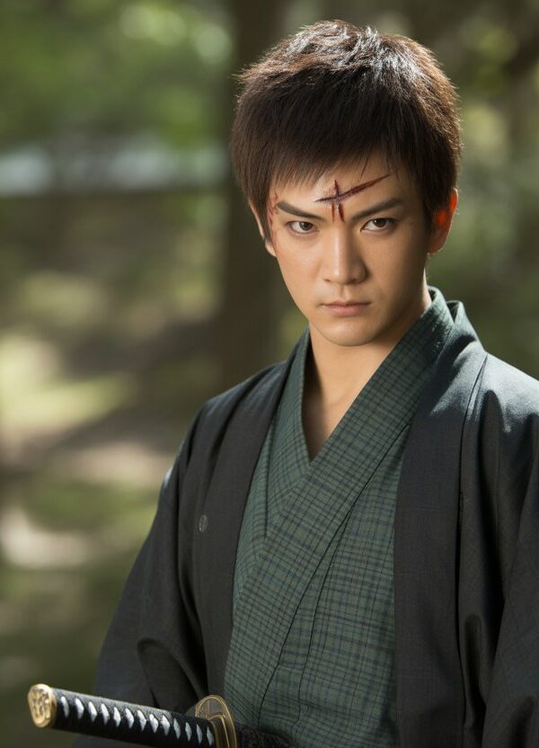 Um jovem guerreiro japonês com cabelo escuro e uma cicatriz na testa, vestindo um quimono verde e preto, em uma floresta com luz do sol filtrada entre as árvores.