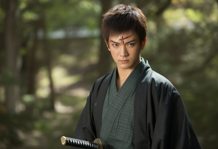 Um jovem guerreiro japonês com cabelo escuro e uma cicatriz na testa, vestindo um quimono verde e preto, em uma floresta com luz do sol filtrada entre as árvores.