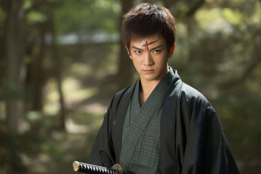 Um jovem guerreiro japonês com cabelo escuro e uma cicatriz na testa, vestindo um quimono verde e preto, em uma floresta com luz do sol filtrada entre as árvores.