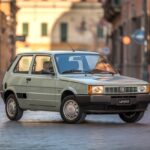 Um carro Fiat Uno clássico estacionado em uma rua de cidade italiana com prédios históricos ao fundo.