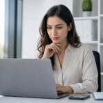 Mulher jovem sentada em uma mesa de escritório moderna, usando um laptop e com expressão pensativa.